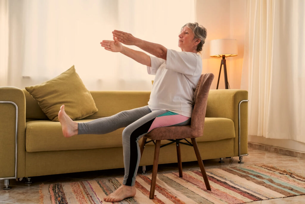 Femme âgée assise au bord d'une chaise dans son salon, une jambe tendue devant elle, le buste incliné vers l'avant.
